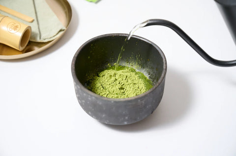A small dark bowl filled with green matcha powder, with hot water being poured into it from a black gooseneck kettle on a white surface.