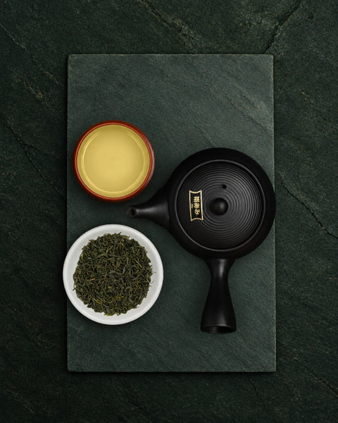 A black ceramic teapot with gold calligraphic detailing on the lid, arranged on a dark slate tray alongside a small white bowl of oolong loose green tea leaves and a small red-rimmed cup of pale yellow tea, photographed from above on a dark green textured surface.