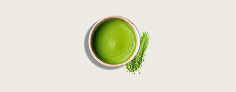 A top-down view of a small bowl filled with bright green matcha tea, with a small pile of loose green matcha powder beside it on a light beige background.