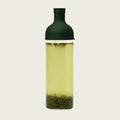 A clear glass bottle with a dark green cap, filled with pale yellow-green tea and showing loose tea leaves at the bottom, set against a solid off-white background.