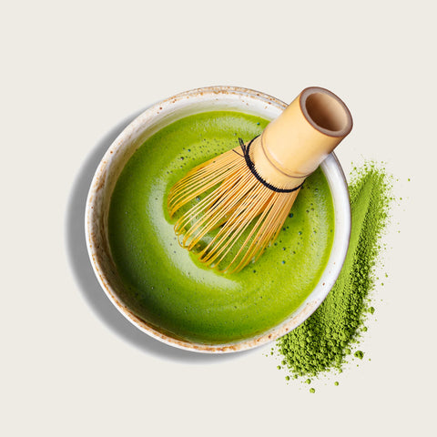 A ceramic bowl filled with bright green matcha tea and topped with a bamboo whisk, with a small pile of green matcha powder beside it on a light beige background.