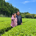 Fujiwara Farmer couple standing in a lush green tea plantation under a bright blue sky, with tall trees in the background.