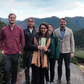 Nio Teas's Team and Satoen Farmers standing outdoors on a path with mountains and greenery in the background. The central person is holding a small booklet or package with Japanese text.