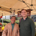 Zenkouen farmer standing under a beige canopy tent at what appears to be a market or outdoor event. A hanging sign behind them includes the partial text 'Org.