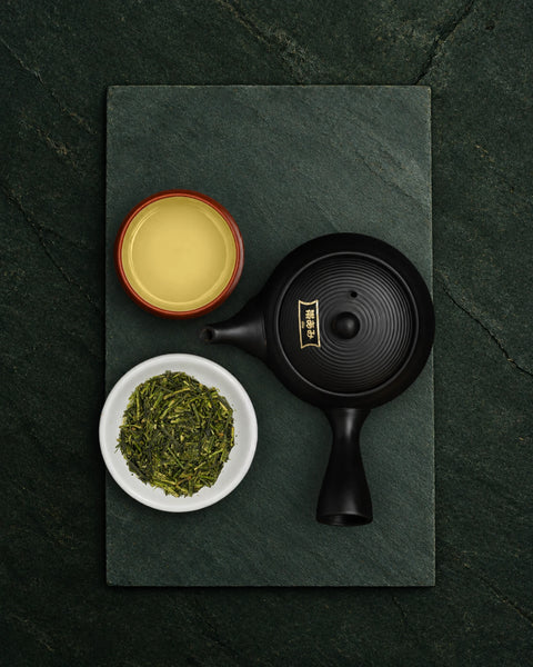 A black ceramic teapot with gold Japanese characters on the lid, displayed on a dark slate tray alongside a small bowl of karigane loose green tea leaves and a cup of brewed yellow tea.
