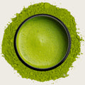 A top-down view of a black matcha bowl filled with bright green matcha foam, surrounded by a larger ring of pure matcha powder on a light off-white background.