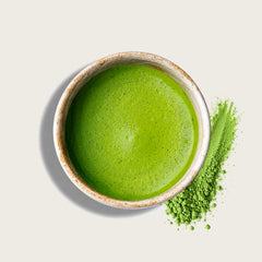 A ceramic bowl filled with bright green matcha tea with a small pile of green matcha powder beside it on a light beige background.